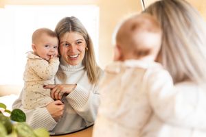 Smiling blonde mother holding her happy baby while looking in a mirror in a bright room.