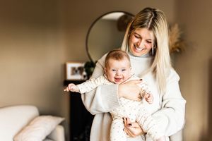 Joyful blonde mother in cream sweater holding her smiling baby in a cozy home setting.
