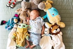Newborn baby sleeping peacefully surrounded by colorful stuffed animals including Baby Yoda and a lion on a green blanket.