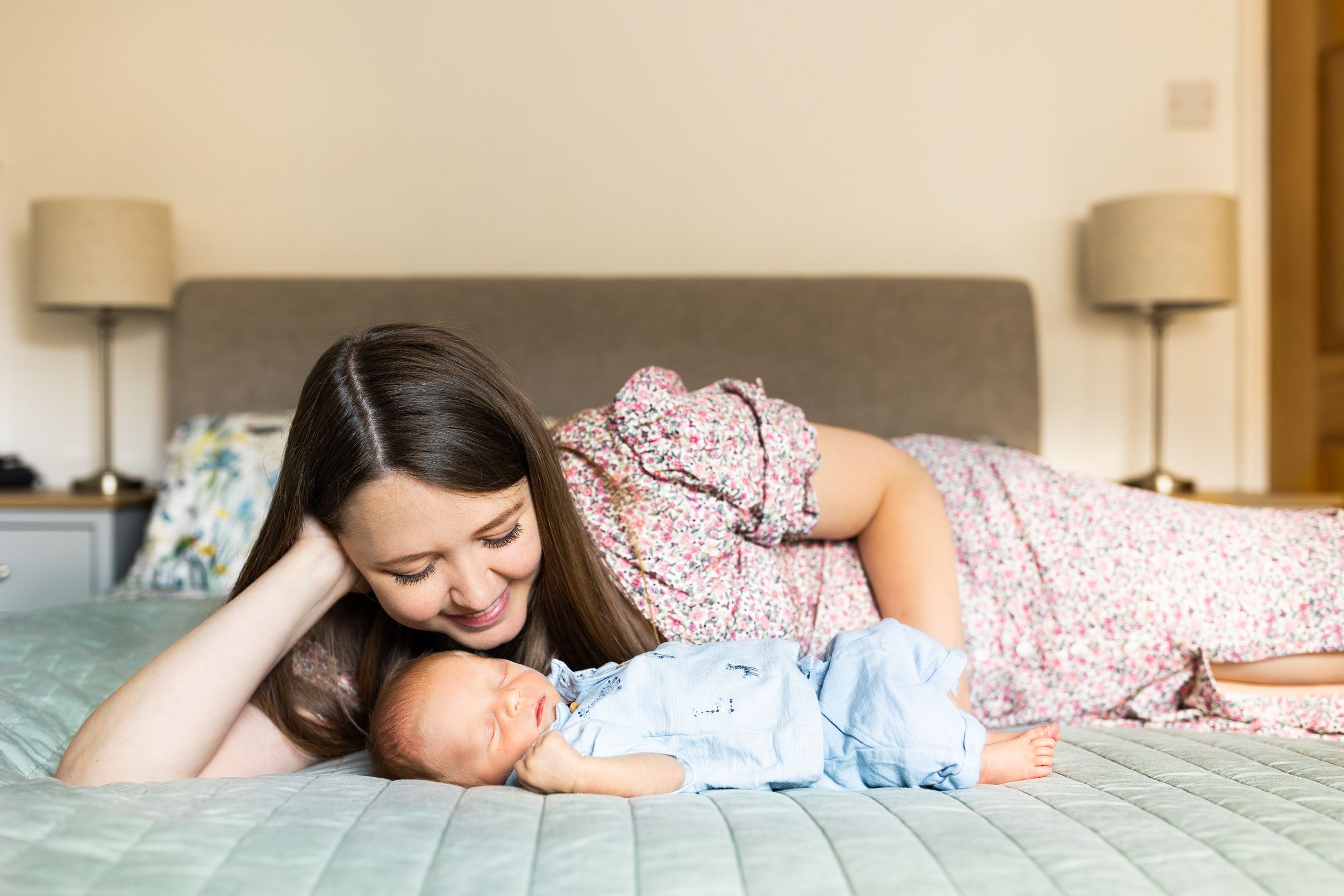 Smiling brunette mother lying on a bed gazing lovingly at her sleeping newborn baby in blue outfit.