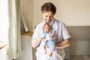 Happy father in striped shirt cradling his newborn baby dressed in blue near a bright window.