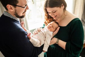 Parents with glasses smiling and cradling their sleeping newborn baby by a bright window.