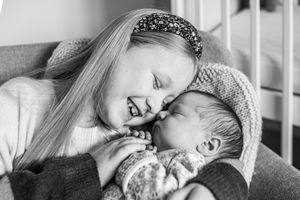 Black and white photo of smiling older sister tenderly holding and nuzzling newborn sibling.