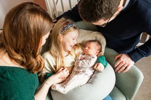 Family with mother, father, young daughter, and newborn baby gathered in cozy armchair.