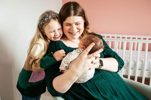 A joyful mother in green dress holds her newborn while her laughing toddler daughter hugs her.