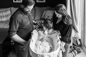 Black and white family portrait of parents and child gazing at newborn in a bassinet.