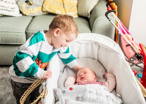 A young boy leans over a bassinet, gently touching his sleeping newborn sibling.