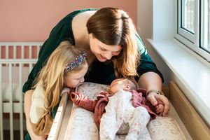 Mother and young daughter lovingly looking at newborn baby in crib by window.