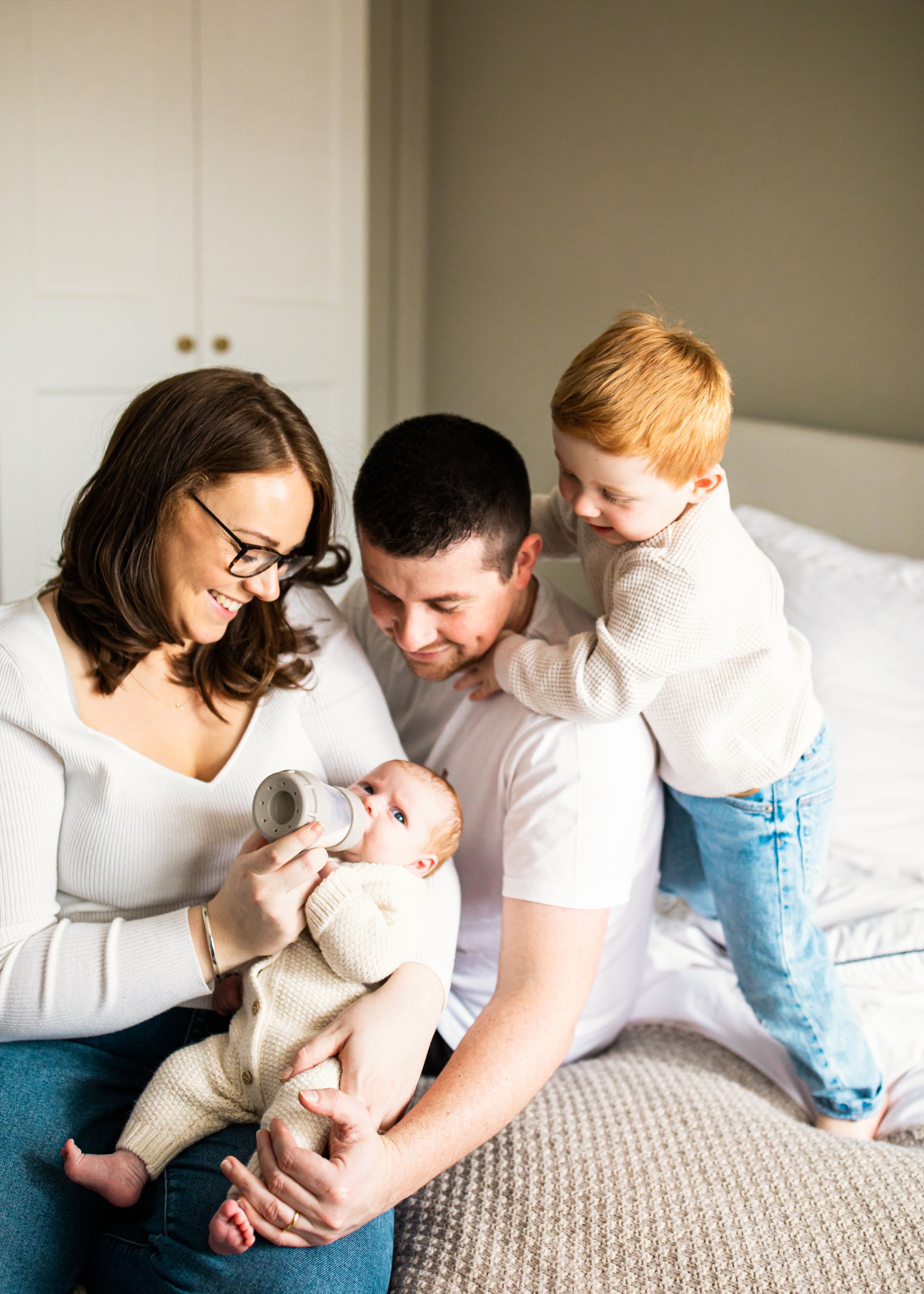 Happy family on a bed as parents smile at their baby being bottle-fed while toddler watches.