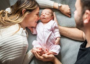 Loving parents leaning over their sleeping newborn baby in pink outfit on grey blanket.