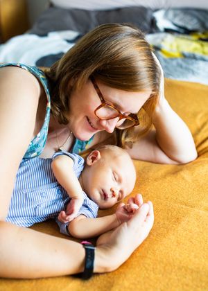 Smiling mother with glasses lying on yellow bed gazing at her sleeping baby in blue romper.
