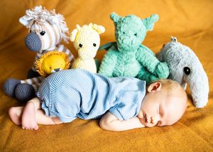 Sleeping baby in blue striped romper surrounded by colorful stuffed animal toys on yellow background.