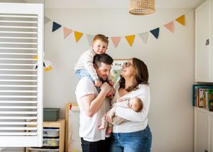 Smiling parents in a colorful nursery, dad holding toddler on shoulders, mom cradling newborn.