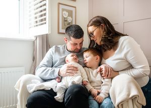 A loving family of four sits together by a window, parents gazing at their newborn baby.