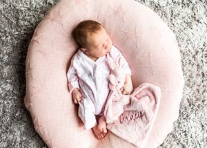 Sleeping newborn baby in pink outfit on round pink knit cushion with personalized blanket.