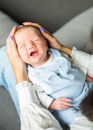 Mother gently cradling yawning newborn baby in light blue outfit on grey sofa.