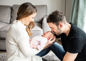 New parents sitting on floor smiling at their sleeping newborn baby girl in pink outfit.