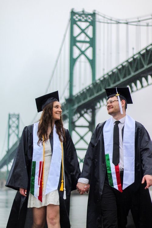 Graduates in caps and gowns hold hands in front of a green suspension bridge.