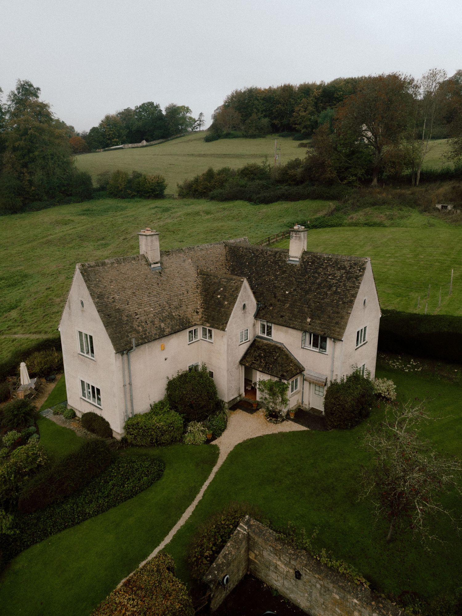Aerial views of a charming white cottage with thatched roof nestled in rolling green countryside hills in England.