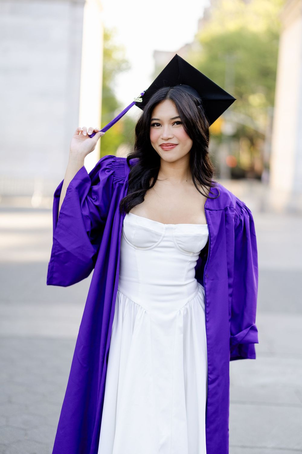 Graduate wearing purple graduation gown and cap with white dress smiling in outdoor photo session.