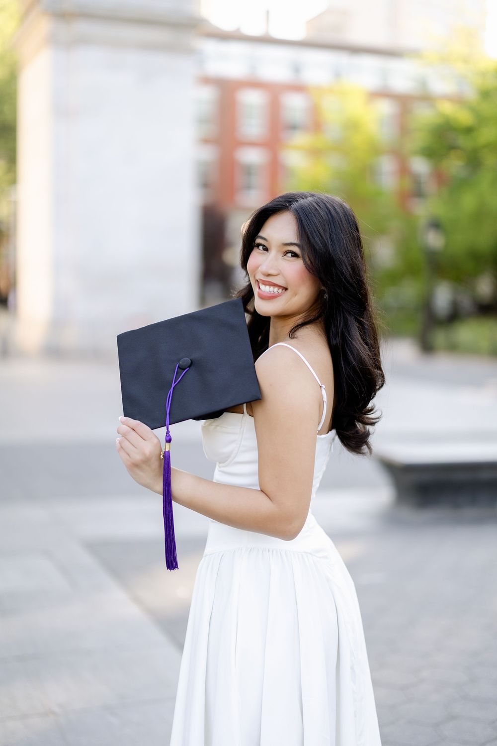 A graduate holds up a black graduation cap while wearing a white dress on a sunny day.