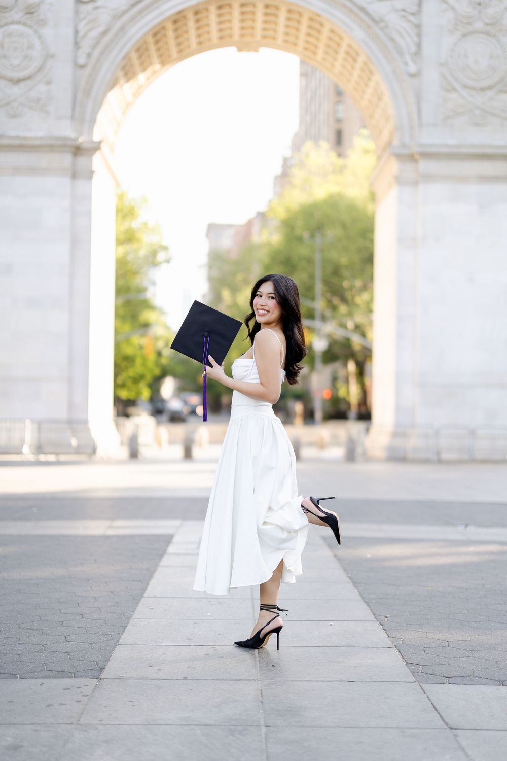 A woman in a white dress and black heels poses gracefully under a stone archway in an urban setting.