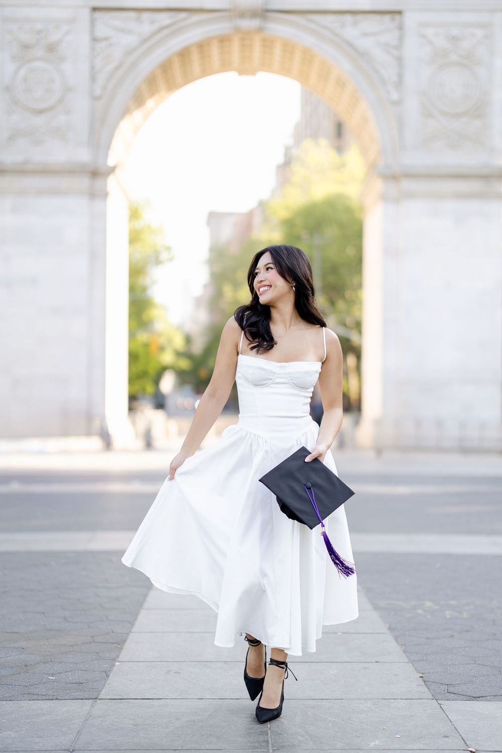 A woman in a flowing white dress and black heels twirls gracefully under a stone archway in an outdoor setting.