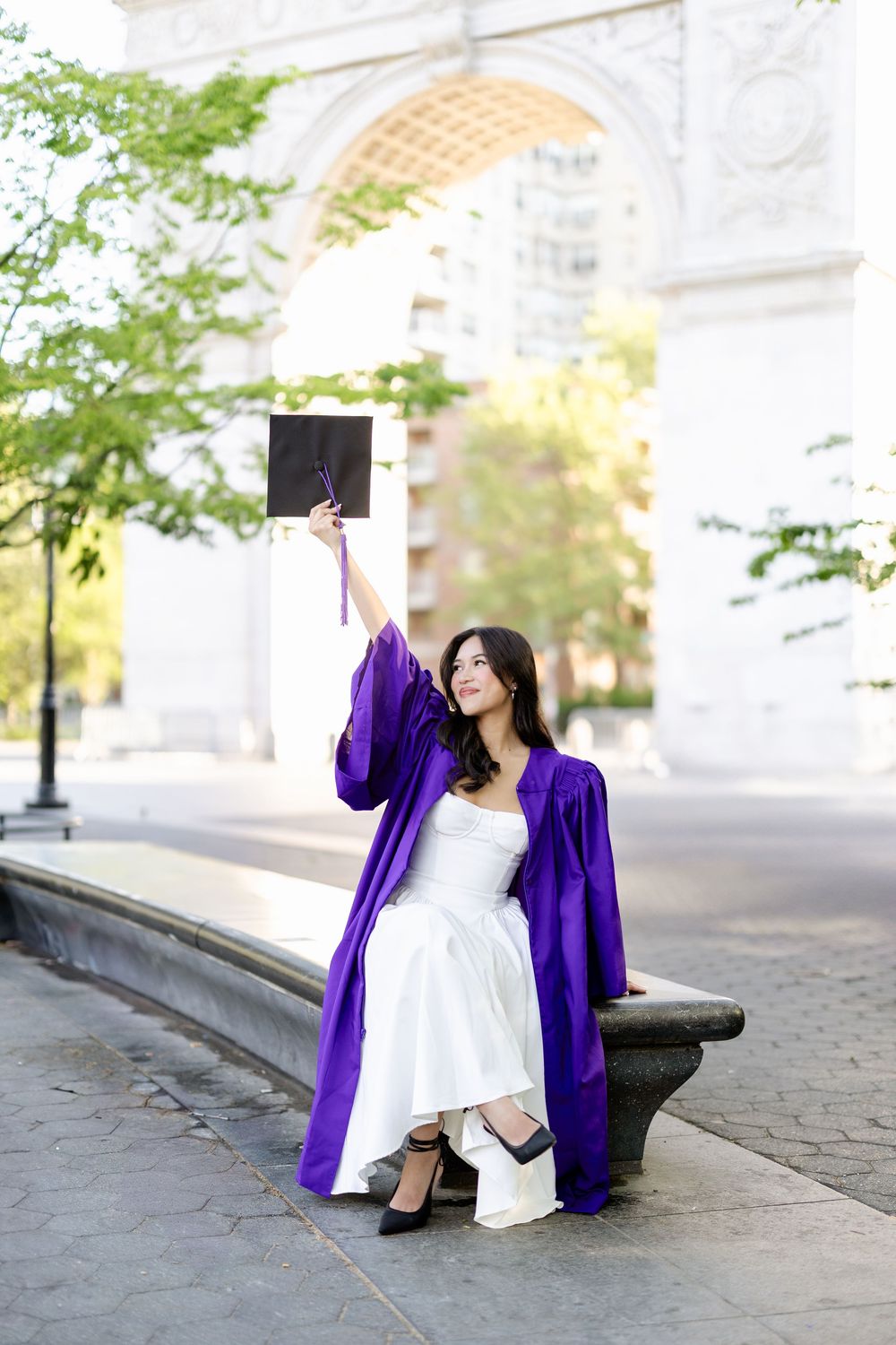 Graduate in purple graduation gown and white dress sits thoughtfully on stone steps during outdoor photo session.