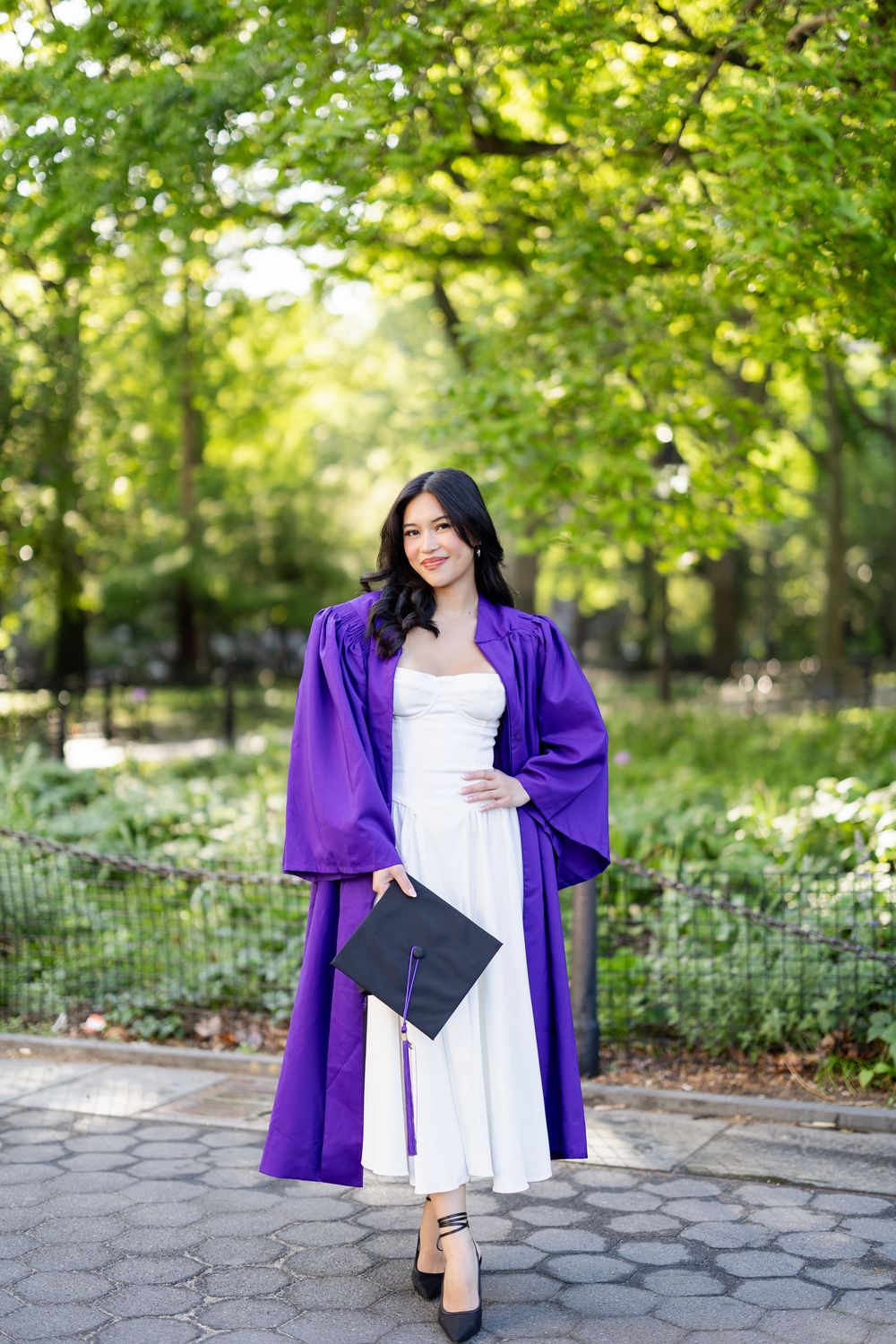 Graduate in white dress and purple graduation gown holds diploma while posing in park setting.
