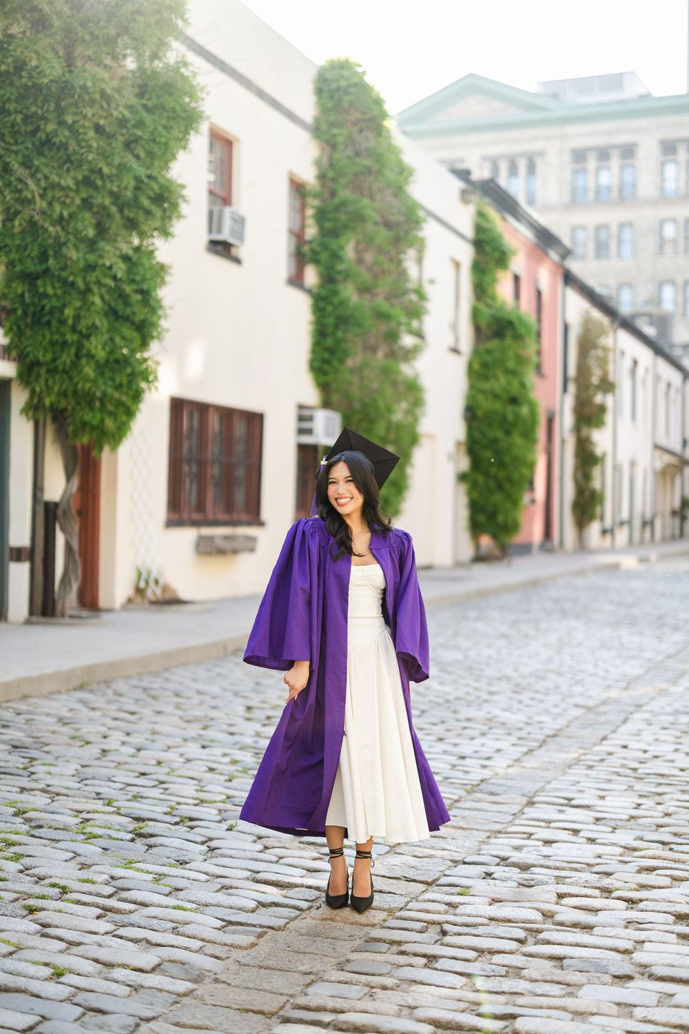 A graduate poses on a cobblestone street wearing a white dress and purple graduation gown.