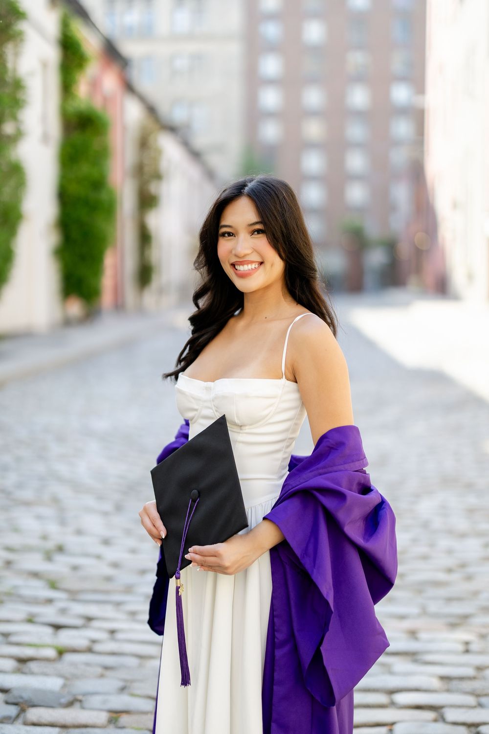A graduate in a white sundress and purple stole poses outdoors against a brick building backdrop.