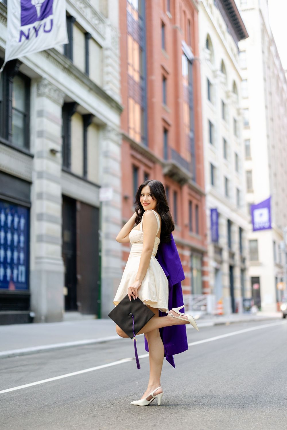 A person in a black dress and white boots crouches on a city street with NYU buildings in the background.