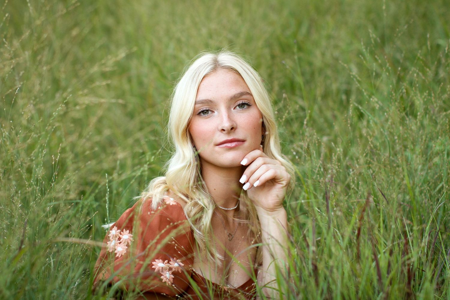 A blonde young woman surrounded by tall green grasses smiles softly for the camera during her Kansas City senior photo session.