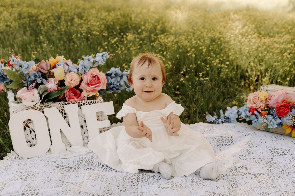 A baby in a white dress lies on a lace blanket surrounded by colorful flowers in a dreamy outdoor setting.