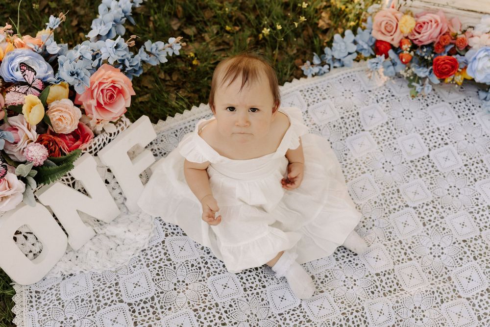 Small child in white dress sitting on delicate lace blanket with pink and blue floral arrangements.