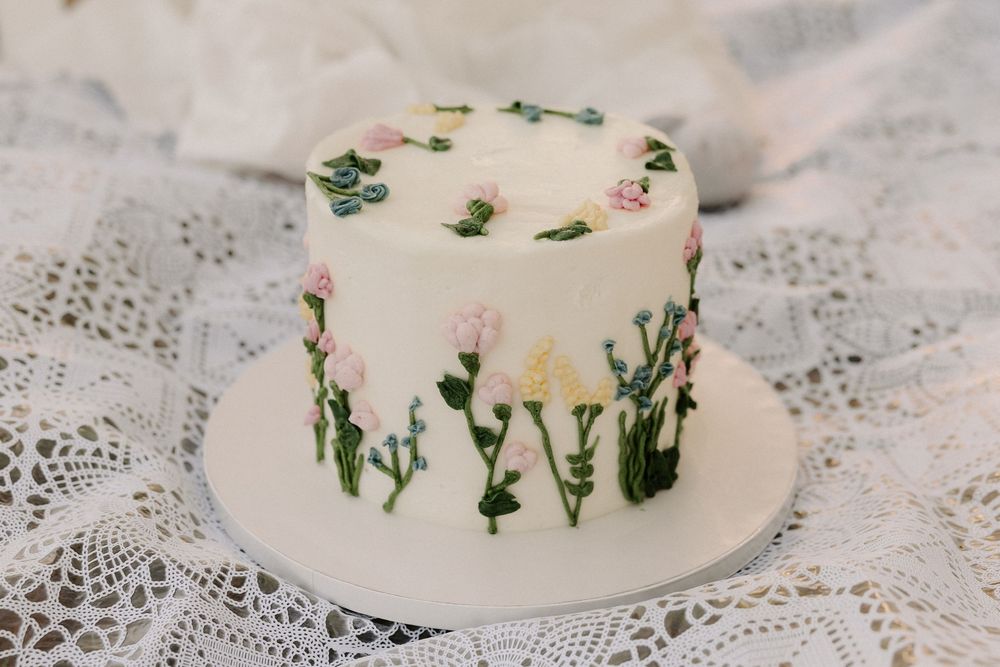 A white buttercream cake decorated with delicate pink and green floral details sits on a lace tablecloth.
