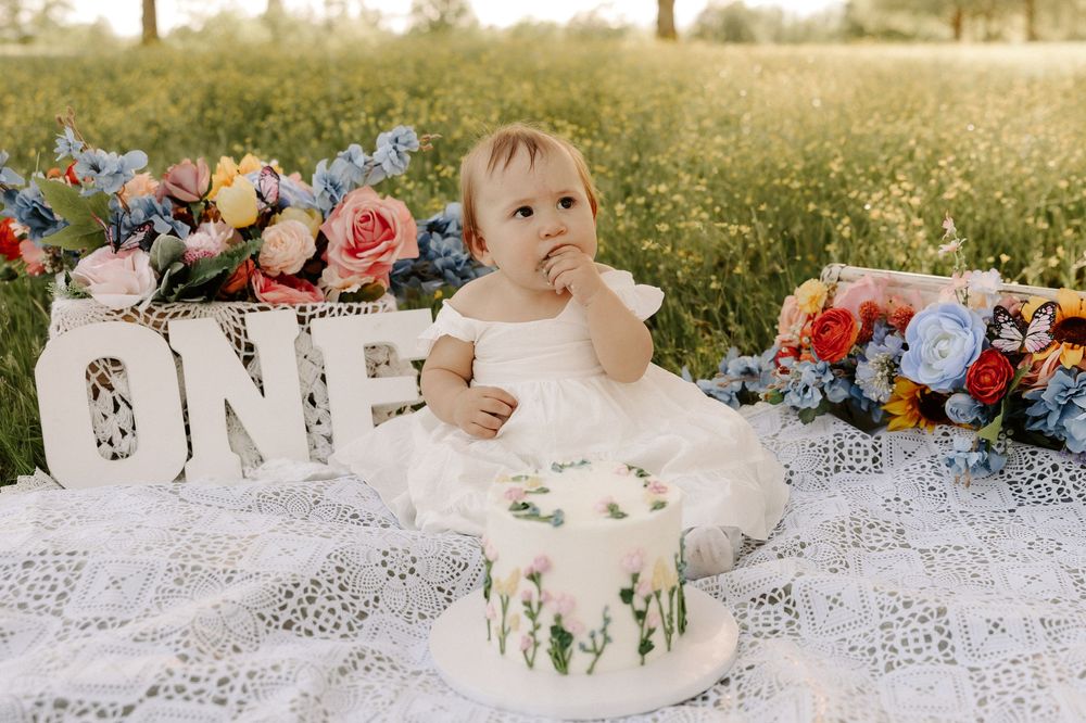 A white cake with floral decorations sits beside decorative 'ONE' letters on a lace blanket outdoors.