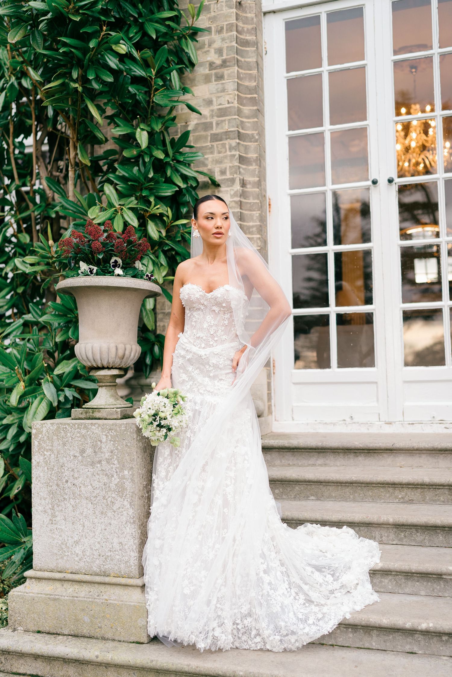 Bride in elegant lace wedding gown poses gracefully on stone steps with greenery and windows in background.