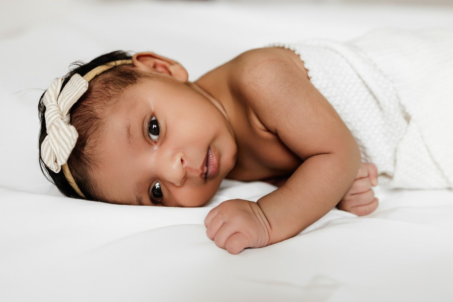 A newborn baby lays on the bed covered by a white swaddle