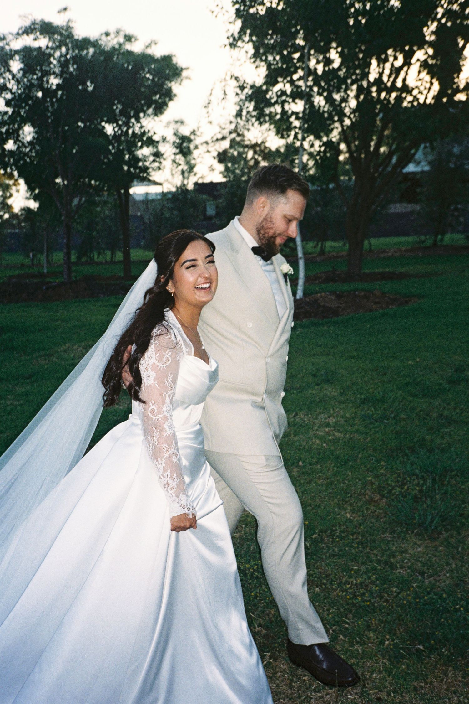 A bride and groom share candid laughing moments in a grassy outdoor setting at sunset.