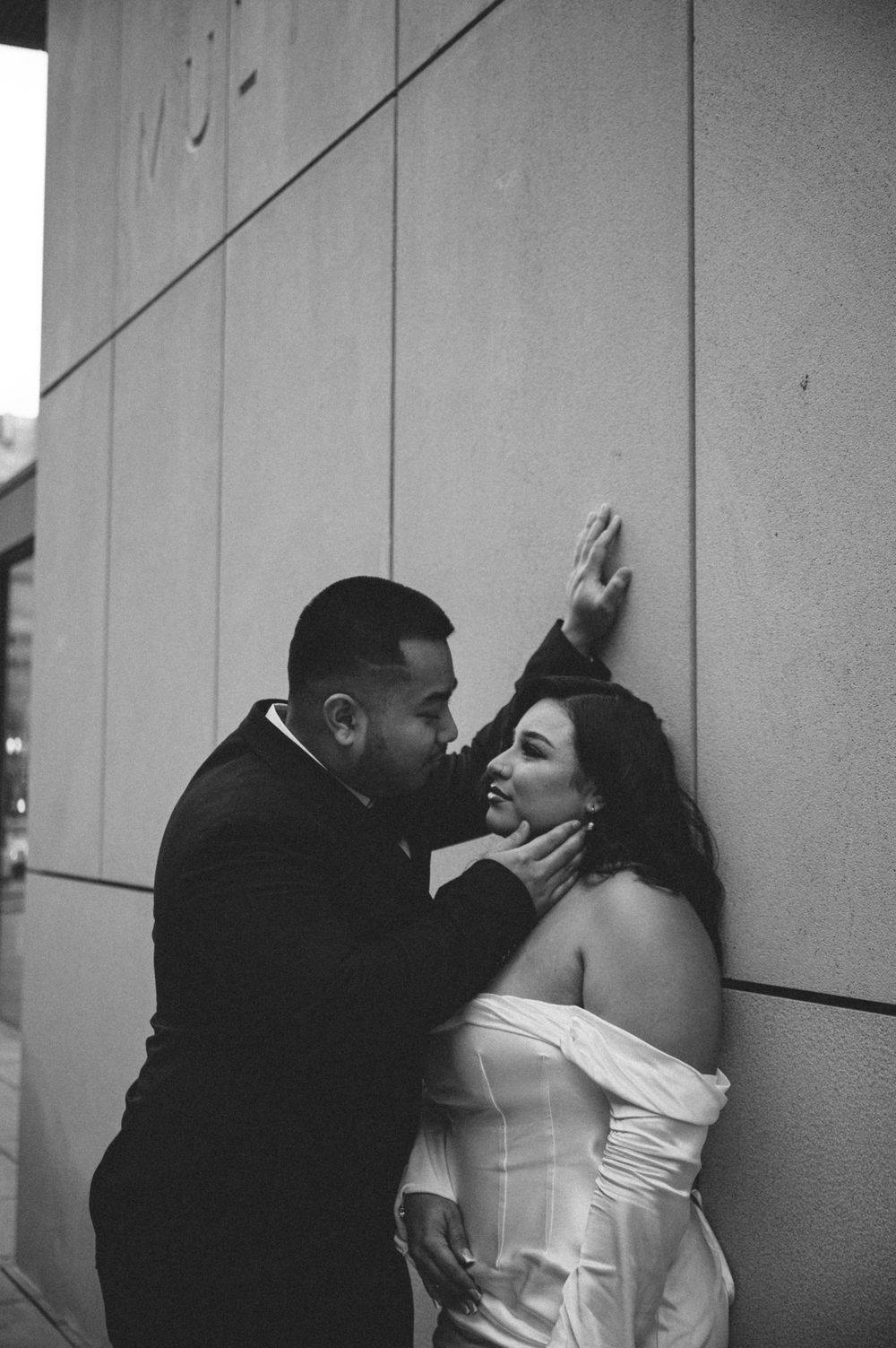 A romantic black and white portrait of a couple in formal attire sharing an intimate moment against a wall.