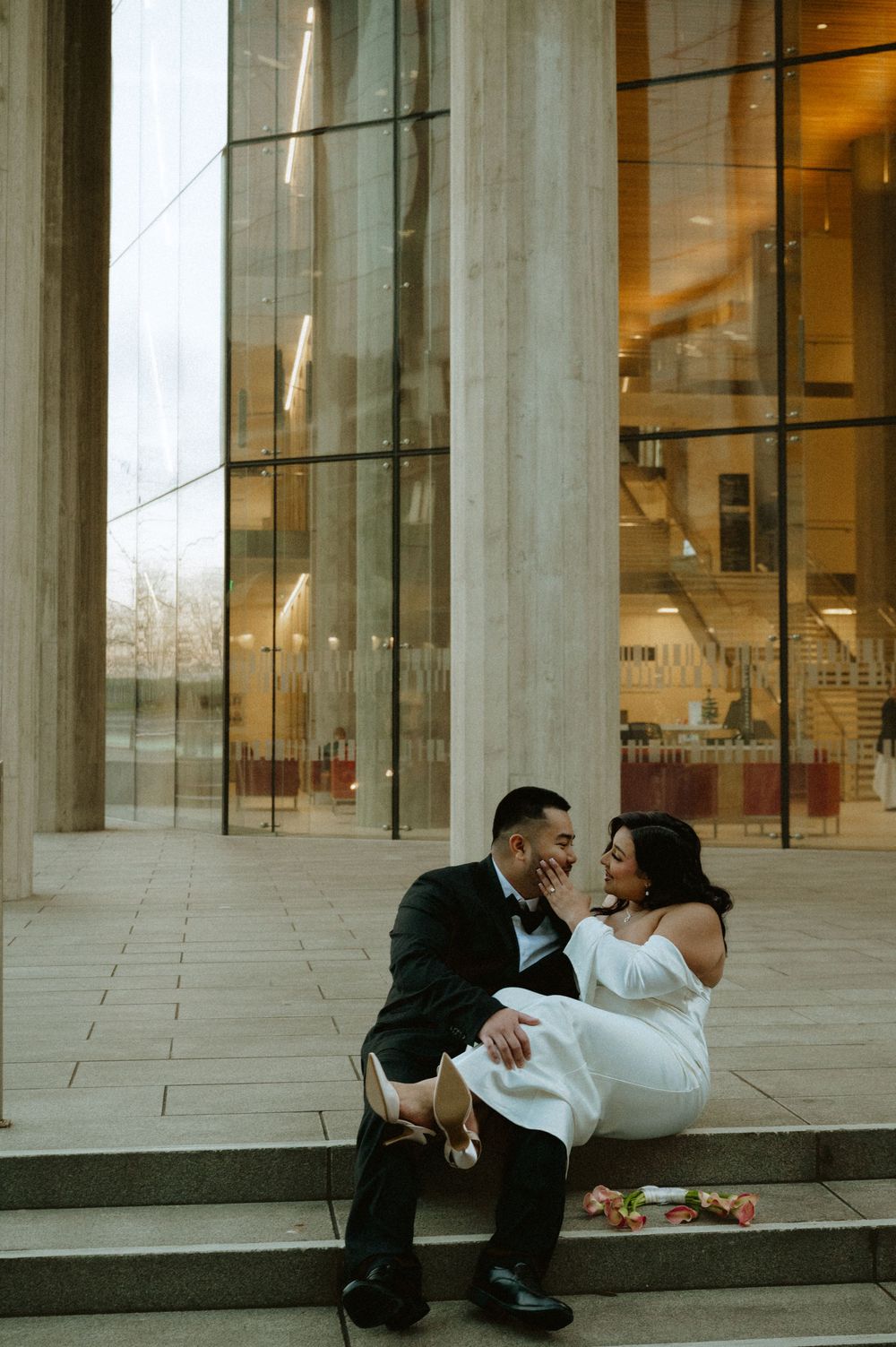 A couple in wedding attire sits together on stone steps outside a modern glass building.