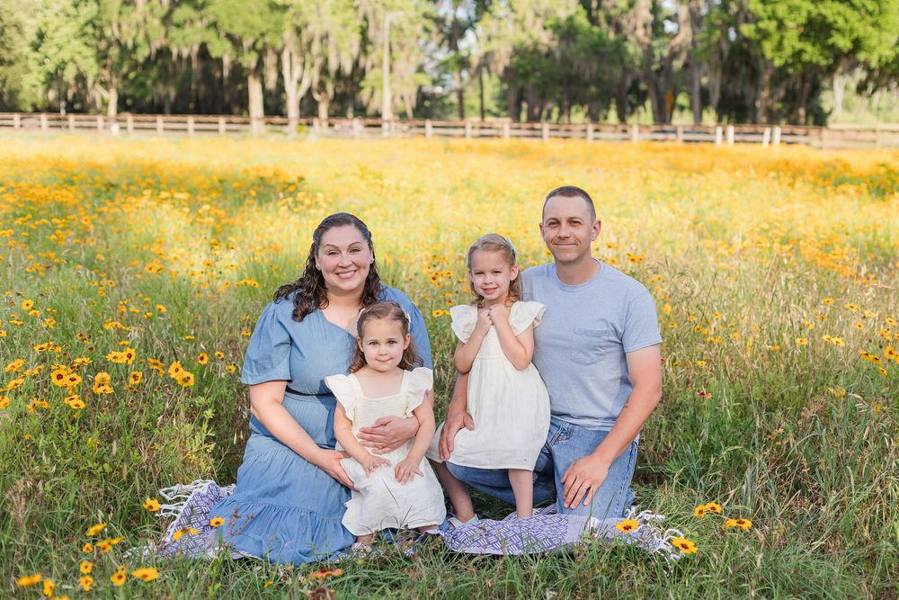 Family sits together on blanket in field of yellow wildflowers with oak trees in background.