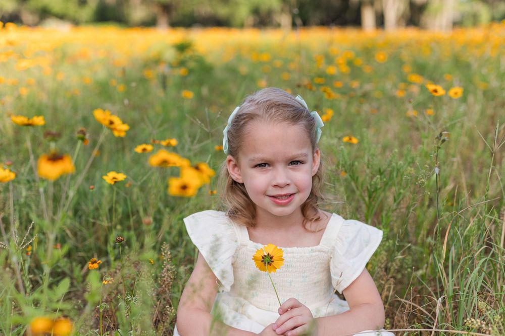 Two children in matching white dresses stand together in a meadow of bright yellow flowers.