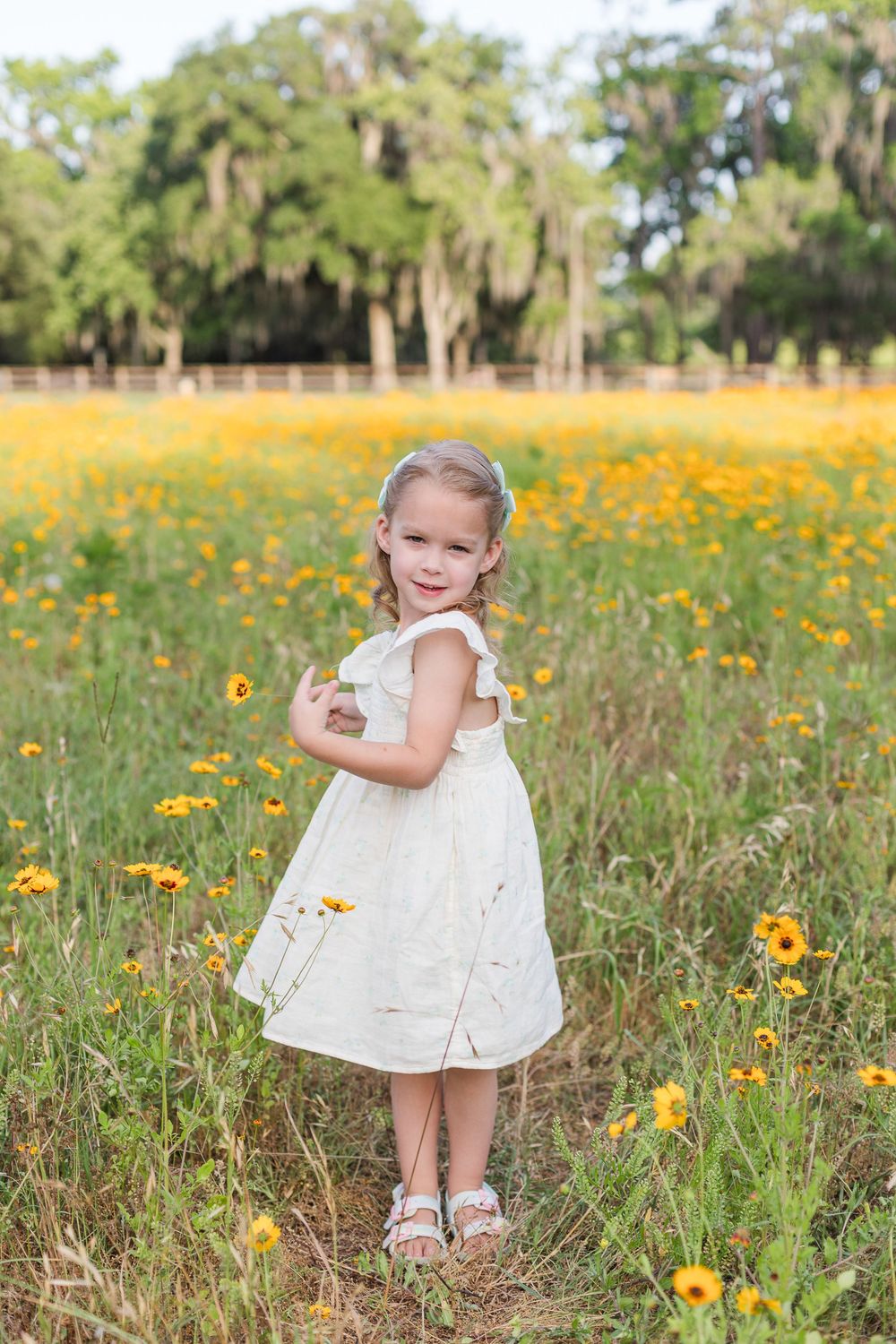 A white eyelet dress stands out against a field of yellow wildflowers and Spanish moss-draped trees in the background.