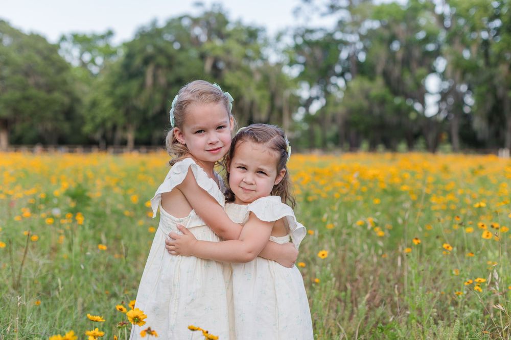 Two children embrace while wearing white dresses among yellow wildflowers in a natural outdoor setting.