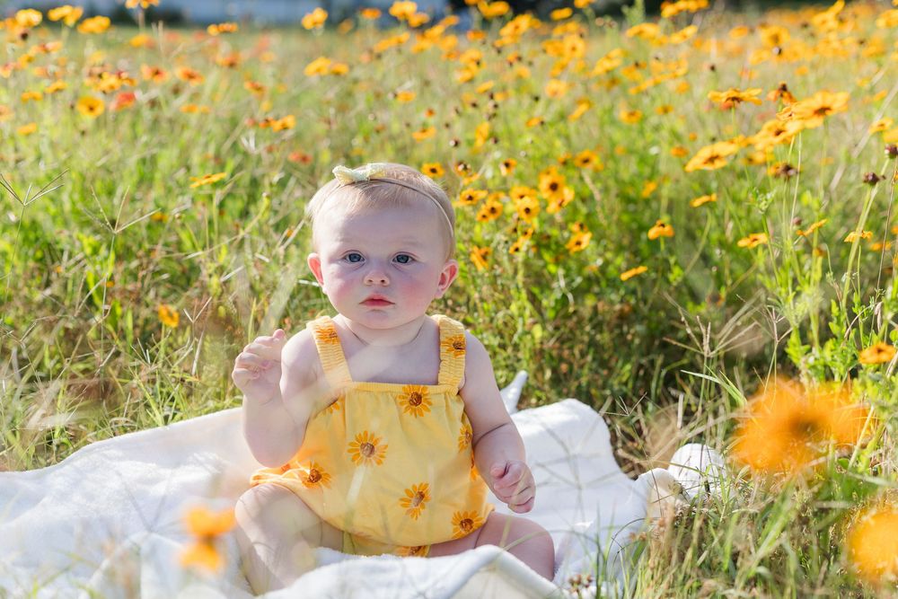 A child sits among vibrant orange wildflowers in a sunny meadow on a white blanket during a summer photo session.