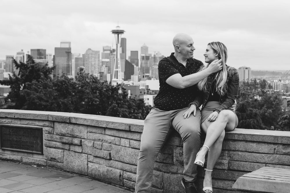Couple embraces against stone wall backdrop with city skyline in background.