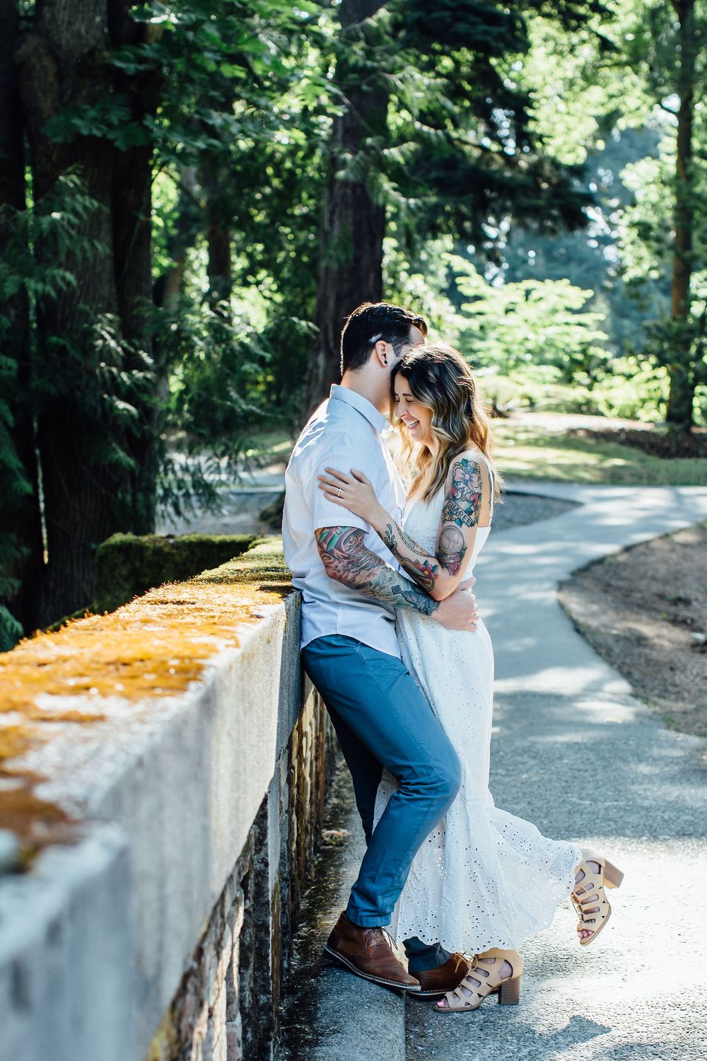 Romantic couple poses along stone wall on gravel path surrounded by lush greenery.