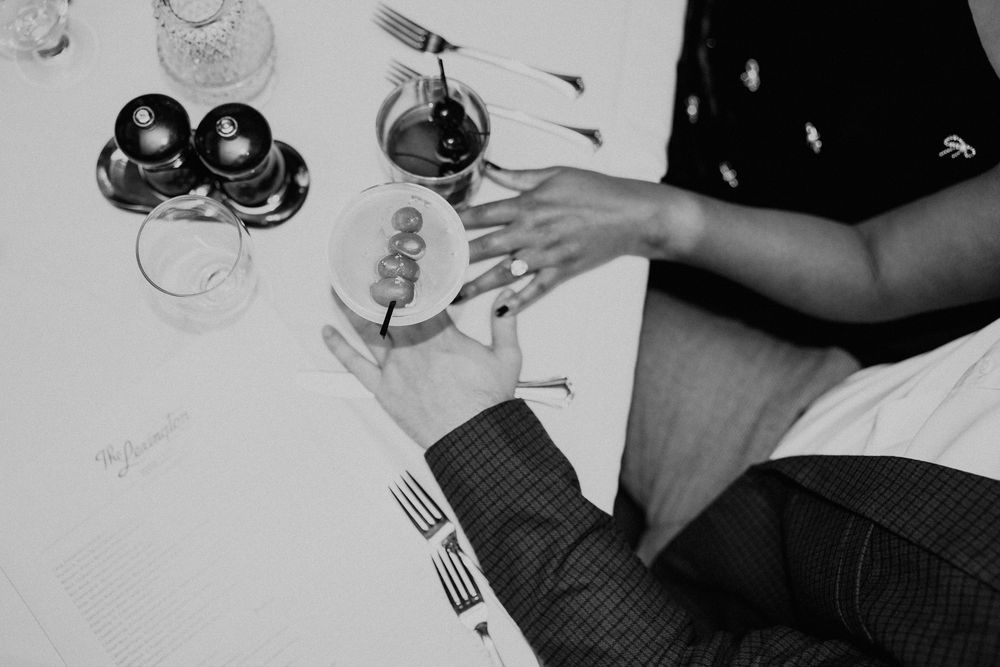 Black and white photo of hands holding wine glasses and forks at a dinner table setting.